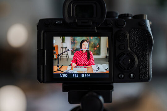 camera screen shows a digital artist in a striped pink shirt at a desk with sketchbooks and a tablet, framed by a professional recording setup in a modern studio.