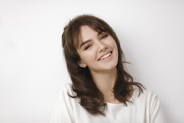  Portrait of a cheerful young Caucasian woman with wavy brunette hair, wearing a casual white top and smiling warmly. Ideal for lifestyle, beauty, or promotional concepts. 