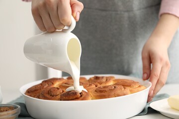 Woman adding frosting onto freshly baked cinnamon rolls at table indoors, closeup