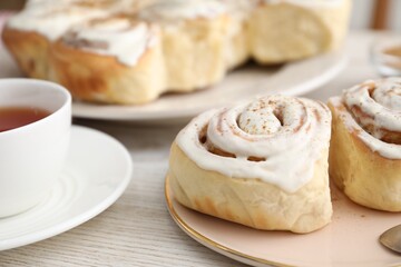 Delicious frosted cinnamon rolls and cup of tea on light wooden table, closeup