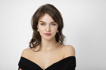 Naklejka premium A stunning brunette woman with gold earrings poses in a black off-shoulder dress against a minimalist white background. Perfect for style, beauty, and fashion photography concepts. 