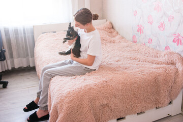 A young woman sits on a plush pink bed, gently holding a small, dark-colored rabbit. The setting appears to be a bedroom with soft lighting.
