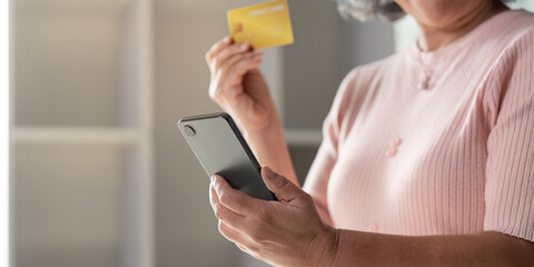 Elderly woman focusing on her smartphone while holding a credit card for online shopping in a bright home setting