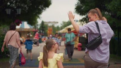 Woman in purple shirt and girl with Down syndrome in yellow dress walk hand in hand at entrance to amusement park. Scene conveys excitement, anticipation, and shared experiences