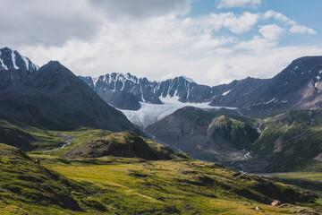 Dramatic scenery in alpine valley with creek among green hills and rocks with view to rocky pointy peak, large snow-capped peaked top, mountain range and big glacier tongue under clouds in blue sky.