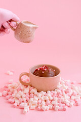 hand with creamer and chocolate bomb in a ceramic mug sprinkled with marshmallows isolated on a pink background