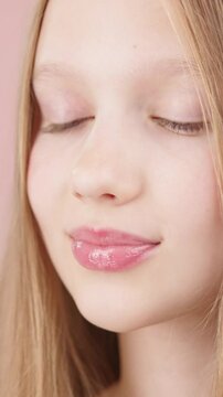 VERTICAL VIDEO. Pretty young girl applying lip gloss. Caucasian blonde girl gen Alpha using make-up cosmetics, posing in studio on pink background. Concept of advertising cosmetics for teens.