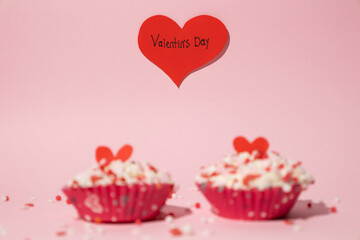 Two out of focus cupcakes decorated with colorful candies. Behind them there is a red heart with a written message that says Valentine's Day. On a pink background. Concept of love and affection