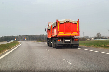 An orange grain dump truck with a semi-trailer transports grain along the road from a farm. Transportation of grain harvest. Copy space for text, agricultural © HENADZY