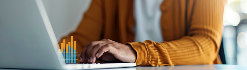A business woman works on her laptop while sitting at her desk. She is wearing a yellow sweater and has her hands on the keyboard. There is a graph on the screen.