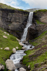 Waterfall on the mountain in black stone that forms a river in a green valley with stones
