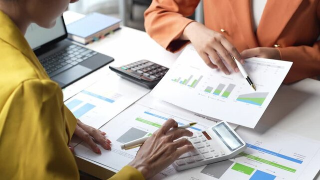 Two women are looking at a table with papers and a calculator. They are discussing numbers and graphs