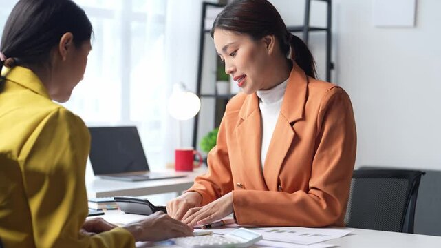 Two women are sitting at a desk, one of them is wearing a yellow jacket. They are talking to each other and one of them is pointing at something on the table