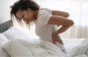 A woman in a light shirt sits on her bed with a pained expression, holding her lower back as she struggles with discomfort in a well-lit bedroom
