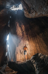 Adventurer exploring a massive sunlit cave with dramatic rock formations and natural light beams, wearing shorts and standing barefoot on a rocky ledge.