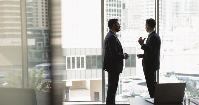 Silhouettes of two businessmen dressed in formal suits stand against panoramic window with skyscraper cityscape view, discuss partnership opportunity, joint project completion, and future investments
