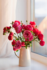 On the table by the window there is a white vase with a bouquet of pink garden roses.