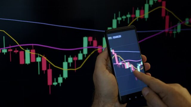 Trader or investor monitoring or looking stock market or crypto market graphics or charts on phone and PC. Man&rsquo;s hand holding phone with graphics in front of PS screen.