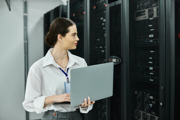 A woman in a white shirt works hard on a laptop in a server room, ensuring system reliability.