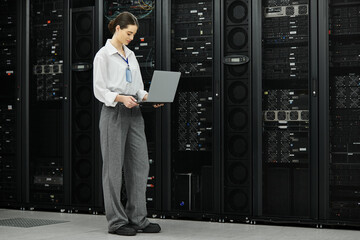 A woman in a white shirt examines a laptop in a contemporary server room filled with hardware.