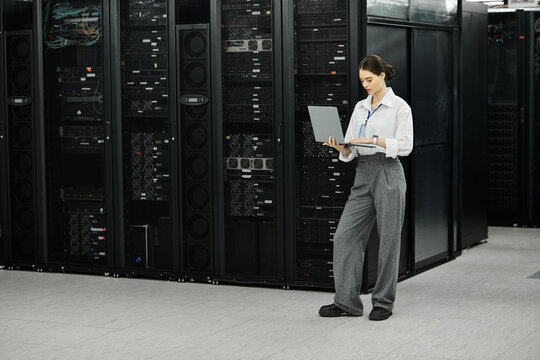 A woman in a white shirt and gray trousers manages server hardware in a data center. - Powered by Adobe