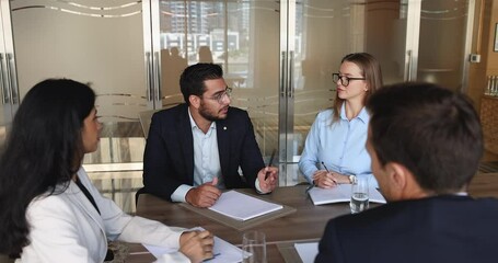 Young Arabian businessman conduct business meeting seated at conference table with clients or staff members, express point, explain joint project details, make speech, looks confident. Seminar event