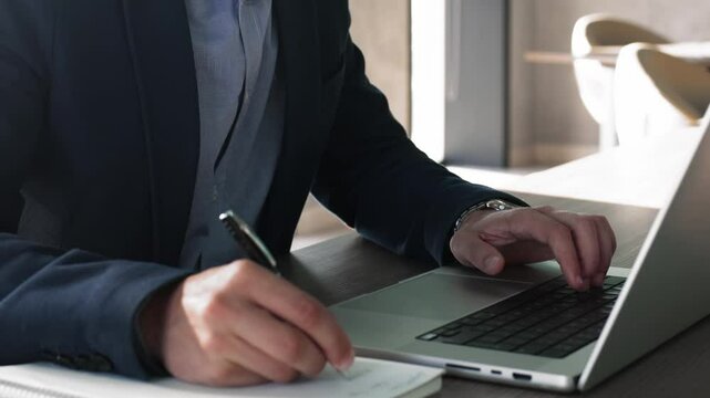 Businessman in suit working on laptop seated at desk, holding pen, making notes, jotting important information in personal organizer, planning strategy, e-learning, gains new knowledge, close up view