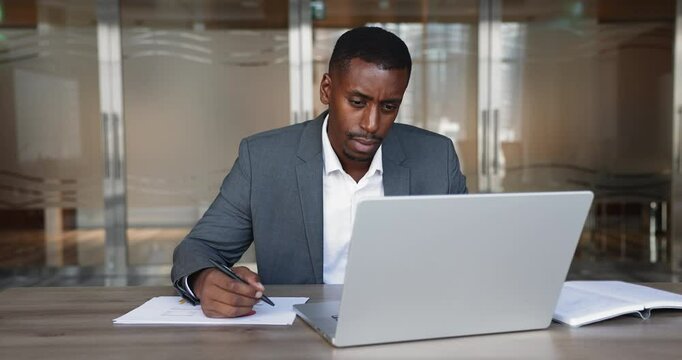 Serious African businessman dressed suit working at desk in office, researching information, making notes on paper and using laptop, check reports, websurfing info on internet, jotting important data
