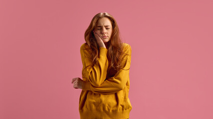 Young red-haired woman suffering from exhausting toothache, holding hand on her cheek on pink...