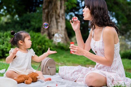 Little toddler catching soap bubbles and having picnic with mother in park