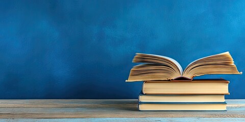 Fototapeta premium Open hardback book atop neatly stacked books on wooden table against solid blue backdrop ample copy space ideal for education and back to school themes