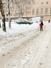 Fototapeta premium A person walks on a snow-covered street next to parked cars in a city during winter, highlighting urban winter challenges