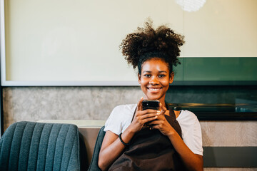 Close-up portrait of a black female barista and coffee shop owner using a mobile phone. Working in uniform smiling communicating joyfully. Woman's relaxation is apparent.