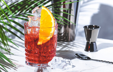 Mezcal negroni, alcoholic cocktail drink with agave spirit, red bitters, sweet vermouth, ice and orange. Marble table, white walls, palm leaves with shadow