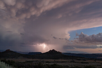 A forked bolt of lightning strikes from a dramatic sky filled with pink and grey clouds above the desert landscape of Hop Valley in Southern Utah, USA on a summer evening.