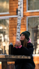 Relaxed young woman in warm winter clothes drinking hot tea outdoors in the backyard of her country house. Nice woman holding a mug with a drink, dreaming, admiring the winter landscape, time for