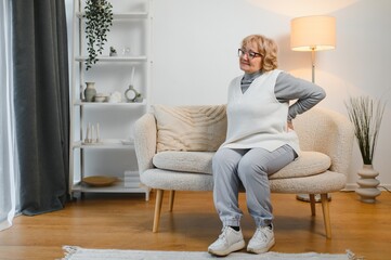 Fototapeta premium Senior woman having backache and touching her back in living room at home