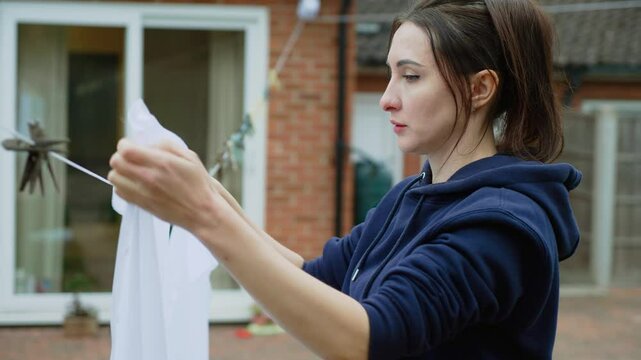 Woman clipping white fabric on outdoor clothesline. Female organizing laundry with focus. Housewife completing drying task in backyard. Person handling household chores attentively