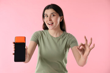 Happy young woman with payment terminal showing OK gesture on pink background