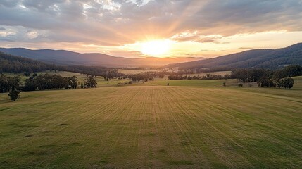 Fototapeta premium A vast agricultural field stretches under a breathtaking sunset, illuminating the sky with vibrant colors. Distant mountains frame the horizon, creating a serene countryside atmosphere