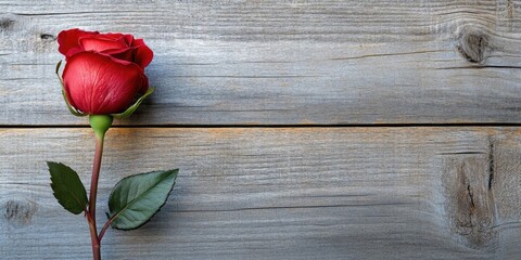Single dark red rose bud positioned left on weathered gray wooden background, creating ideal negative space for text, with green leaves accentuating the rose.