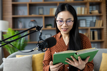 Asian woman with glasses recording podcast at home, reading book. Engaged in creative broadcasting. Relaxed indoor setting, communicating ideas, sharing knowledge.