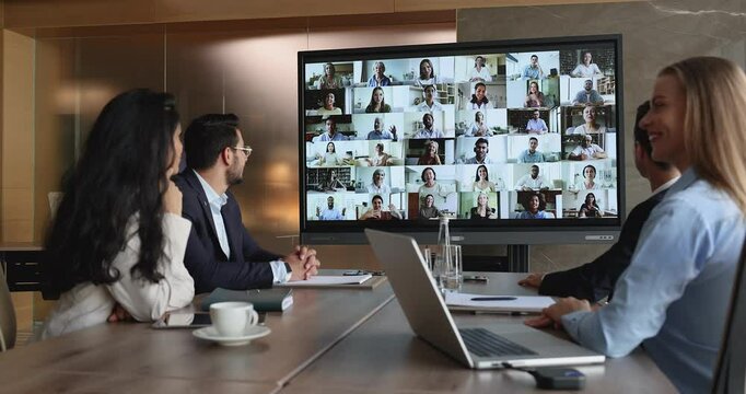 Modern technology usage for remote, digital communication. Group of diverse businesspeople engaged in virtual meeting event seated at table, looking at screen displayed lot of people engaged in talk - Powered by Adobe