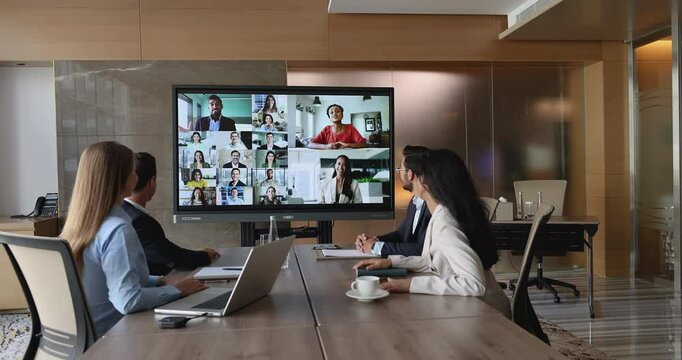 Group of diverse businesspeople participate in video conference seated at table in modern meeting room, interacting with colleagues via videocall app. Global team collaboration, digital communication