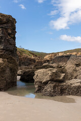 Rocky beach landscape with sandy shores, tide pools, and cliffs under a bright blue sky with scattered clouds.
