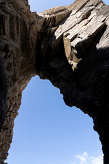 Natural rock arch viewed from below, highlighting rugged textures and blue sky framed by towering cliff walls.