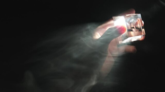 A slow-motion close-up of a male hand holding a transparent glass object toward a beam of light in a smoky atmosphere, illuminated by a low-key dramatic spotlight