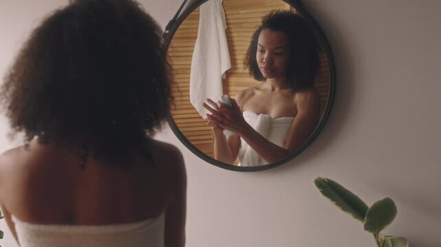 Back view of young Biracial lady in towel looking at mirror reflection and spraying antiperspirant after shower, getting ready for work or school, body care and beauty