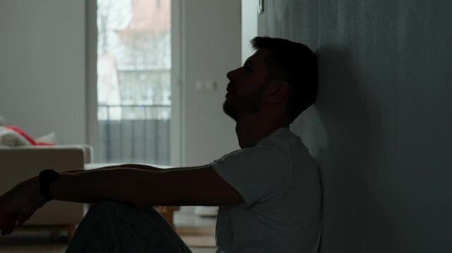Depressed young man sitting on floor in dark room. Silhouette of young male suffering from loneliness and anxiety, covering his face with hands. Person with mental disorder