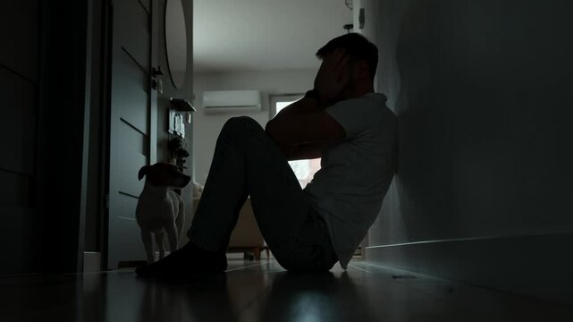 Depressed young man sitting on floor in dark room. Silhouette of young male suffering from loneliness and anxiety, covering his face with hands. Person with mental disorder